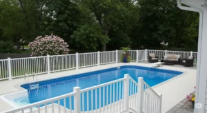 A high-quality, professional photo showcasing a recently installed pool fence and railing project in Buffalo, NY. The image features KD Fence & Deck's signature craftsmanship, with pristine white vinyl fencing and sleek, modern railings surrounding a sparkling blue pool. The backdrop could be a lush, green backyard with blooming flowers or a sophisticated patio setting with comfortable outdoor furniture, conveying a sense of relaxation and safety. The photo is taken from a slightly elevated angle, offering a panoramic view of the entire pool area and showcasing the seamless integration of the fence and railing system. The image conveys a sense of security and peace of mind, emphasizing KD Fence & Deck's expertise in creating beautiful and functional pool enclosures.